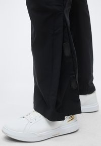 Λευκά δερμάτινα αθλητικά παπούτσια με χρυσό λογότυπο που φοριούνται με μαύρο παντελόνι με πλευρικά κουμπώματα.