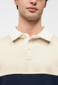 Νεαρός άνδρας που φοράει πουκάμισο με γιακά σε κρεμ και ναυτικό μπλε, εμφανίζοντας το πάνω μέρος του στήθους και το κάτω μέρος του προσώπου σε απλό λευκό φόντο.