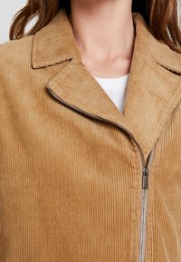 Beżowa kurtka z korduroyu z wyraźnym kołnierzem, wyrazistą pionową fakturą żebrowania oraz srebrnym zamkiem błyskawicznym na froncie.