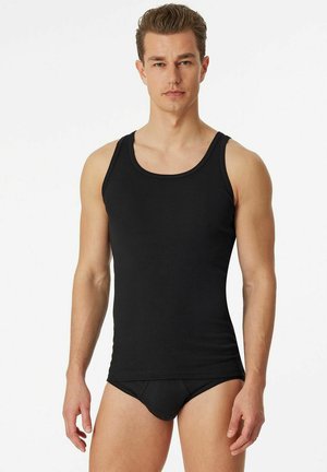 Young man wearing a fitted black tank top and black briefs, standing against a plain white background.