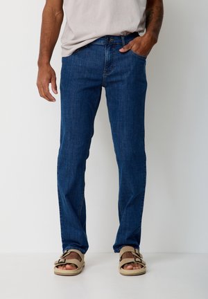 Man wearing blue jeans, beige sandals, and a light beige t-shirt, standing with one hand in pocket against a plain background.