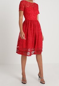 Rotes Spitzenkleid mit kurzen Ärmeln, floralem Muster, tailliertem Oberteil, ausgestelltem Rock und Ausschnitt am Saum. Getragen mit silbernen Riemchenabsätzen.