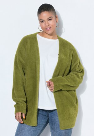 Žena s krátkými vlasy, která nosí zlaté kruhové náušnice, olivově zelený chlupatý kardigan, bílou košili a modré džíny, pózující u bílé zdi.