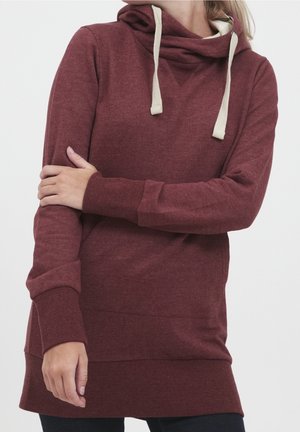 Kobieta w bordowej bluzie z kapturem z kremowymi sznurkami, z założonymi ramionami, widoczna od szyi do połowy uda na jednolitym tle.