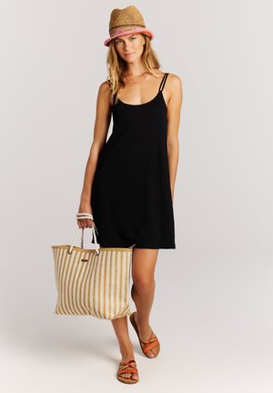 Jeune femme portant une robe noire sans manches, un chapeau de paille, des sandales orange et tenant un grand sac de plage rayé, debout devant un fond uni.