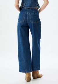 Jeans in denim blu a vita alta e gamba larga, con due tasche posteriori e orlo grezzo. Abbinati a stivali con tacco color beige per un contrasto.