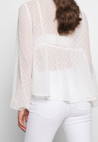 Weiße Bluse mit Punkten und geschichtetem Design, durchscheinender Textur, langen Ärmeln und gerafften Manschetten, kombiniert mit weißen Hosen.