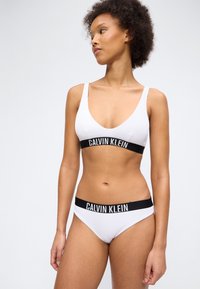 Žena u bijelom Calvin Klein braleteu i odgovarajućim gaćicama s crnim elastičnim trakama s brendom, stoji i gleda u stranu.