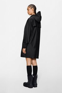 Μαύρο αδιάβροχο παλτό με κουκούλα, με χαλαρή εφαρμογή, μακριά μανίκια και διακριτική πτυχωτή λεπτομέρεια στην πλάτη, σε συνδυασμό με μαύρες μπότες μέχρι το γόνατο.