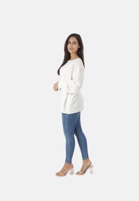 Witte oversized trui met een gestructureerde breisteek, gecombineerd met blauwe skinny jeans en doorzichtige blokhak sandalen, model poseert zijwaarts.
