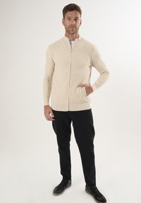 Beige Zip-Up-Cardigan mit geripptem Kragen und Bündchen, mit slim fit. Kombiniert mit schwarzen Jeans und schwarzen Sneakern, vor einem neutralen Hintergrund.