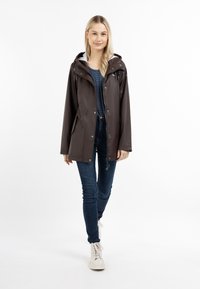 Giacca impermeabile marrone con cappuccio, chiusura frontale a pressione e tasche laterali. Abbinata a una maglietta a righe, jeans e scarpe chiare.