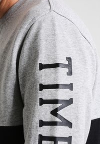 Grijze sweatshirt met zwarte accenten. Heeft een gestructureerde stof en oversized "TIME" letters langs de linkermouw. Contrastkleur aan de onderkant.