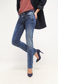 Person som bär blå jeans, marinblå klackskor, en rostfärgad topp och en mörk texturerad jacka, stående med ett ben något upphöjt.