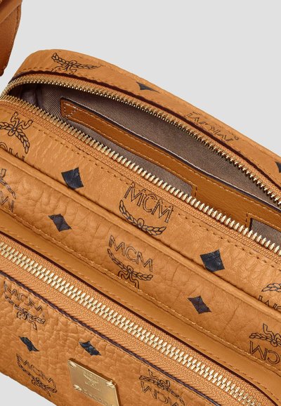 Tan leather bag with black MCM logo patterns, gold zippers, and a small gold metal brand plate on the front pocket.