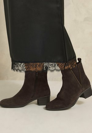 Bottines en daim marron foncé avec talons carrés et fermetures éclair latérales, portées avec une jupe noire à ourlet bordé de dentelle.