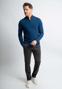 Blauwe gebreide pullover met een halverwege ritssluiting, gecombineerd met donkere broeken en witte sneakers. De pullover heeft lange mouwen en een getailleerde pasvorm.