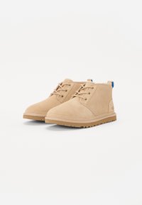Botines de ante beige con diseño de cordones, ojales dorados, suela de goma y acento en el talón azul. Silueta simple y minimalista.