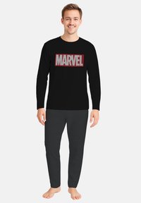 Maglietta nera a maniche lunghe con logo "MARVEL" rosso e bianco, abbinata a pantaloni grigio scuro. L'outfit presenta una vestibilità casual e rilassata.