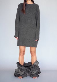 Abito in maglia grigia oversize con polsini e orlo a coste, abbinato a pantaloni in denim grigio a vestibilità larga attorno alle caviglie, piedi nudi visibili.