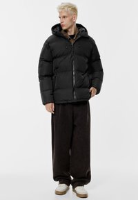 Črna puhovka s kapuco, z zadrgo spredaj in stranskimi žepi. Oblečena z ohlapnimi temnimi hlačami in svetlimi supergami.