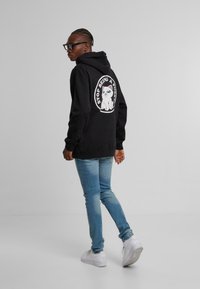 Felpa nera con un design grafico di un gatto e testo sul retro, abbinata a jeans in denim azzurro chiaro e sneakers bianche.