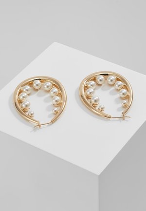 Creolen in Goldton mit gleichmäßig verteilten weißen Perlen im gebogenen Rahmen, auf einem weißen Podest vor grauem Hintergrund präsentiert.