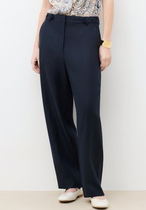 Kobieta ubrana w ciemnogranatowe spodnie o szerokich nogawkach, białe płaskie buty, wzorzysty top i złotą bransoletkę, stojąca na tle białej ściany.