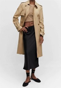 Trench coat beige sopra una maglietta color cammello, abbinato a una gonna nera in satin e ballerine nere. Il cappotto presenta bottoni doppi e una vita cintata.