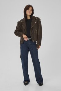 Kvinde iført en mørkebrun oversized læderjakke, sort top, blå viddebukser, sort bælte og sorte spidse sko, stående mod en grå baggrund.