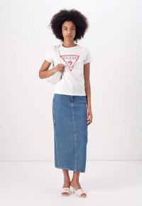 T-shirt bianco a maniche corte con logo grafico, abbinato a una lunga gonna di jeans blu. Gli accessori includono una piccola borsa chiara e scarpe con tacco.