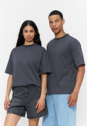 Kobieta w ciemnoszarej oversize koszulce i czarnych krótkich spodenkach dżinsowych stoi obok mężczyzny w ciemnoszarej oversize koszulce i jasnoniebieskich jeansach.