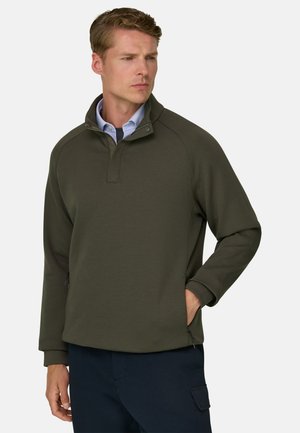 Mand iført en olivengrøn pullover med lynlås i kraven og blå bukser, stående med den ene hånd i lommen mod en ensfarvet baggrund.