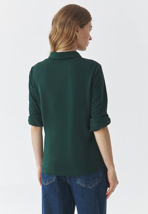 Polo de color verde oscuro con una textura suave, mangas enrolladas y cuello. Combinado con unos vaqueros de denim azul claro.
