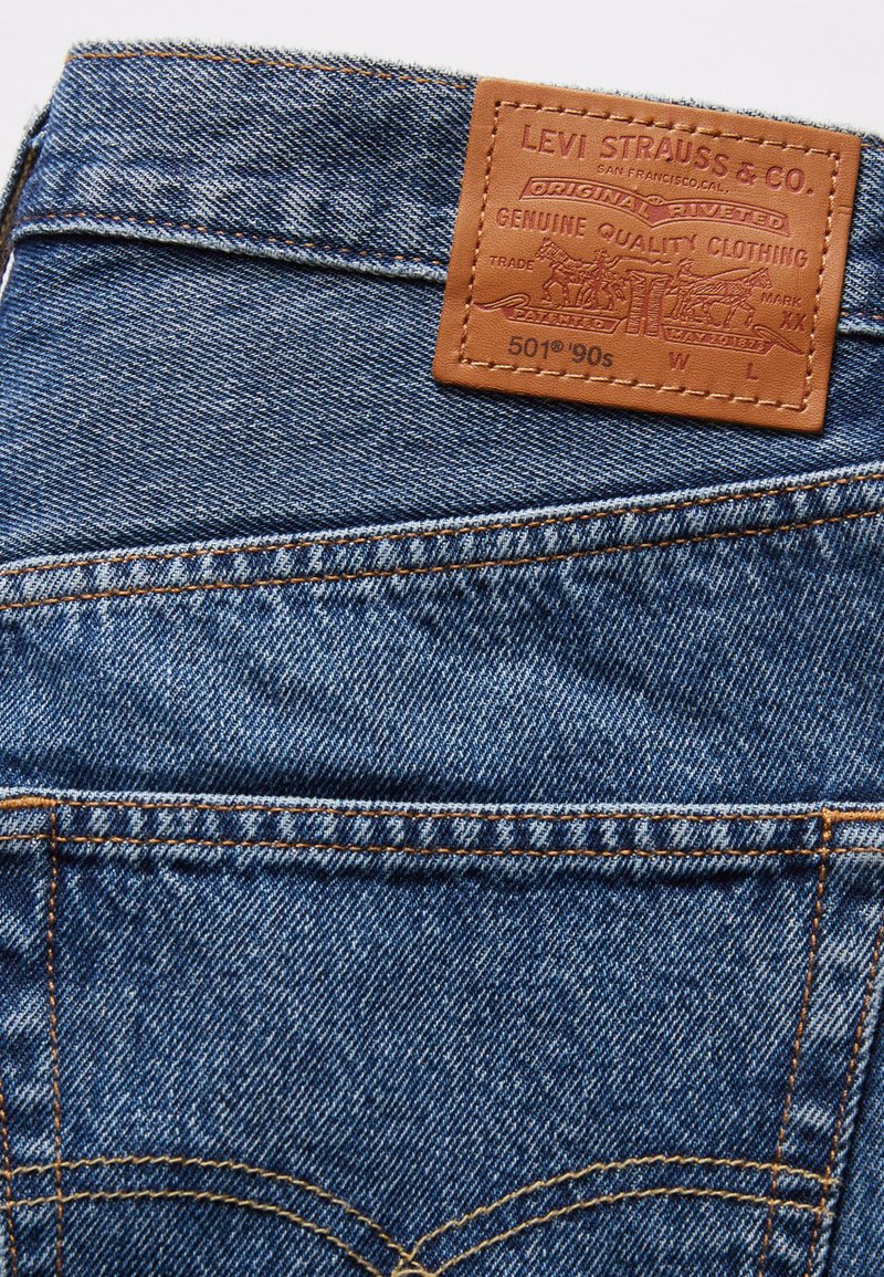 Blaue Jeans aus Denim mit einem braunen Lederetikett, das ein geprägtes Logo und Text zeigt, hervorhebenden Nähten und klassischem Fünf-Taschen-Design.