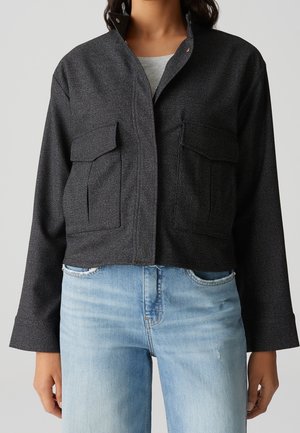 Frau trägt eine dunkelgraue Jacke mit Knöpfen und großen Fronttaschen sowie hellblaue Jeans, dargestellt vom Hals bis zu den Hüften.