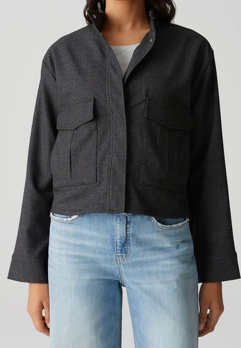 Femme portant une veste boutonnée gris foncé avec de grandes poches avant et un jean bleu clair, vue du cou jusqu’aux hanches.