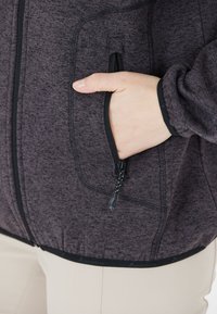 Zip-up jacka i mörklila melerat tyg. Har en sidoficka med svart dragkedja och elastisk midja. Strukturerat mönster genom hela plagget.