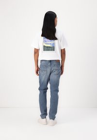 Tricou din bumbac alb cu un grafic pătrat multicolor pe spate, asortat cu blugi din denim albastru deschis și adidași albi.
