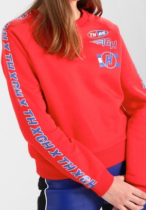 Femme portant un sweat-shirt rouge avec des logos "THXG" bleus et blancs sur les manches et la poitrine, assorti à un pantalon bleu et noir.