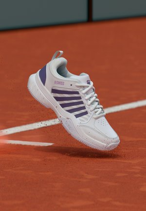 Balts un purpurs K-Swiss sporta apavu pāris, kas spēcīgi sit sarkano māla virsmu tenisa kortā pie baltas robežas līnijas.