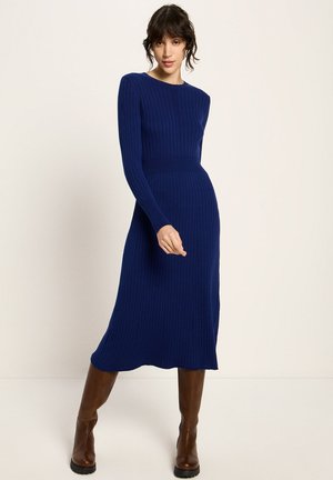 Frau in einem gerippten, langärmligen, marineblauen Midikleid, kombiniert mit braunen kniehohen Stiefeln, steht vor einem schlichten, hellen Hintergrund.