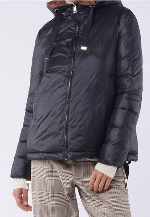 Persoon draagt een marineblauwe donsjas met capuchon en beige geruite broek, staat met handen ontspannen langs het lichaam.