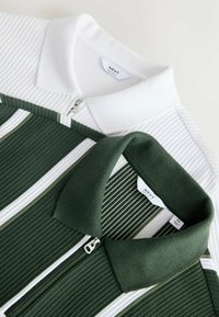 Deux polos en maille côtelée avec fermetures éclair, l'un blanc et l'autre vert foncé avec des rayures blanches et vert clair, posés à plat montrant les cols et les étiquettes.