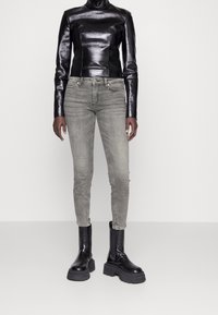 Top largo de cuero negro ajustado, de mangas largas y cuello alto, combinado con jeans gris ajustados y botas negras de tacón ancho. Diseño simple y elegante.