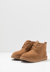 Botas de ante marrón hasta el tobillo con punta redonda, cordones en la parte delantera y suela de goma texturizada; presenta un pequeño logo en el lateral.