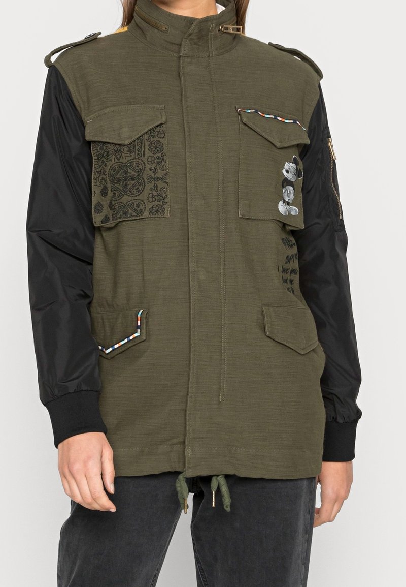 Olive green utility jacket with black sleeves, multiple front pockets, intricate patterned panel, contrasting drawstring hem, and a colorful accent trim.