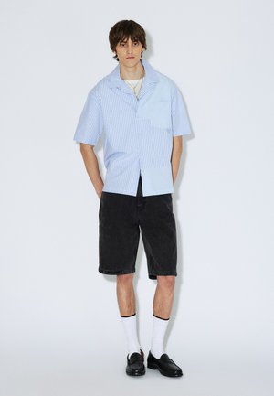 Koszula z krótkim rękawem w jasnoniebieskie i białe pionowe paski, z kieszenią, zestawiona z czarnymi szortami i czarnymi butami oraz białymi skarpetami.