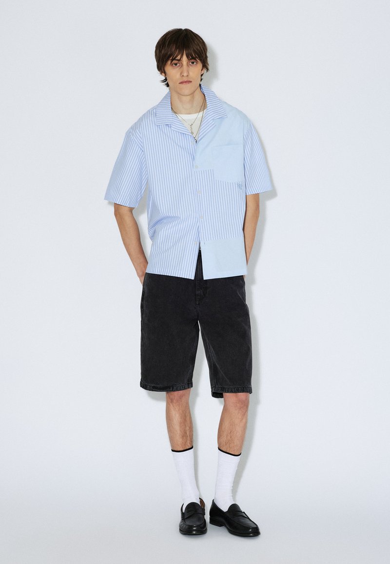 Lyhythihainen paita vaaleansinisissä ja valkoisissa pystysuorissa raidoissa, taskulla, yhdistettynä mustiin shortseihin ja mustiin kenkiin, valkoiset sukat.