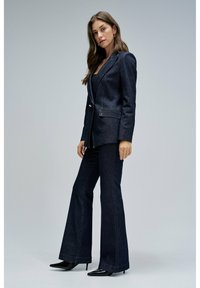 Blazer de denim oscuro con dos bolsillos frontales y cierre de un solo botón, combinado con pantalones de denim oscuro de pierna acampanada. Terminado con tacones negros de punta.